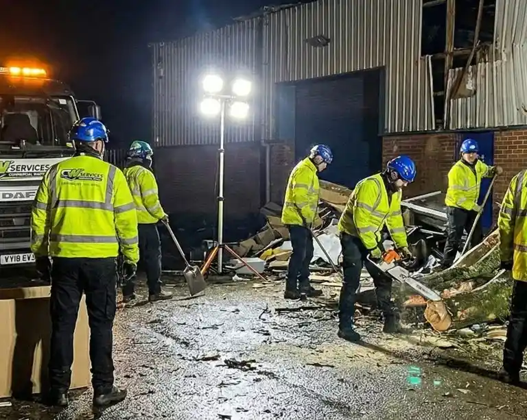 Emergency crew clearing storm damage near an industrial building at night, featuring professional lighting equipment and a service truck.